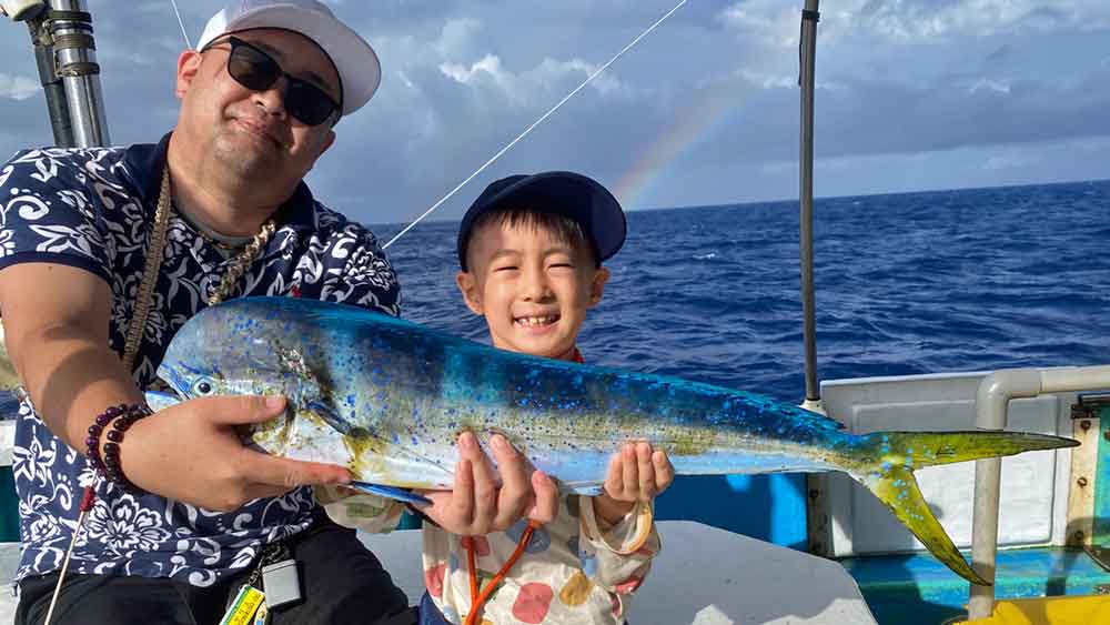 シイラを釣り上げた7才の少年の後ろに虹が見える
