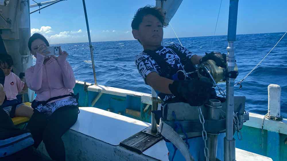 沖縄トローリングでバショウカジキ釣りをしている小学校4年生の少年と後ろで撮影する母